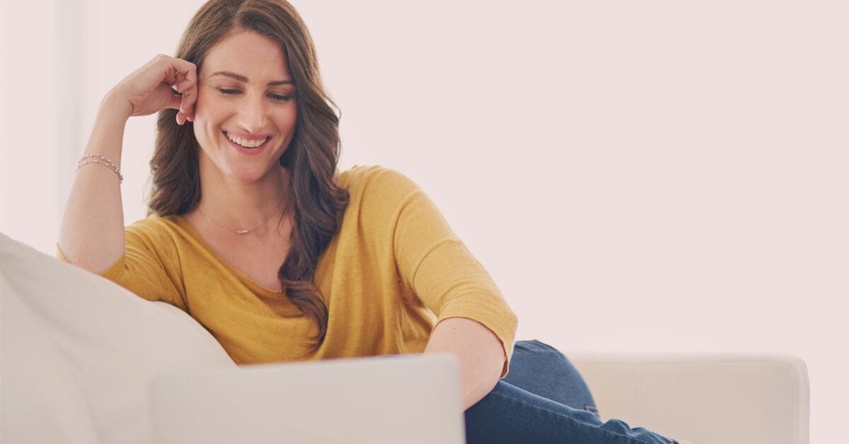 Female entrepreneur smiling while working on laptop, learning how to think like a CEO as an entrepreneur.